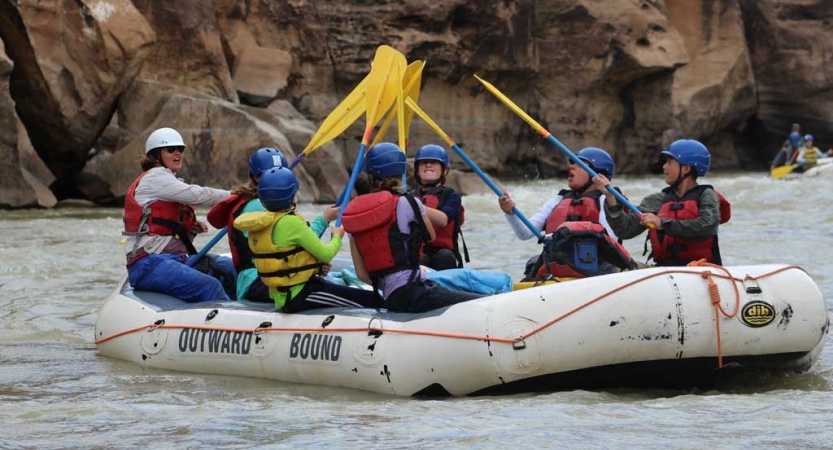 A group of students wearing safety gear sit in a raft and raise their paddles into the air.